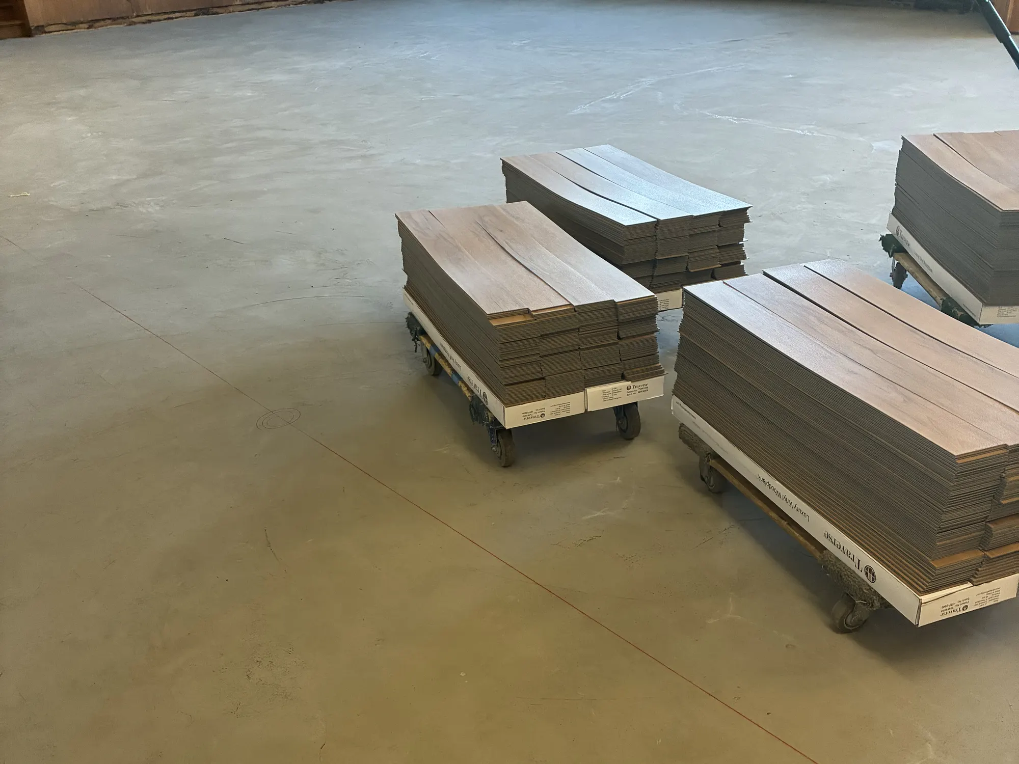 A hall stripped down to a clean, prepared concrete subfloor, with carts of vinyl plank staged for installation
